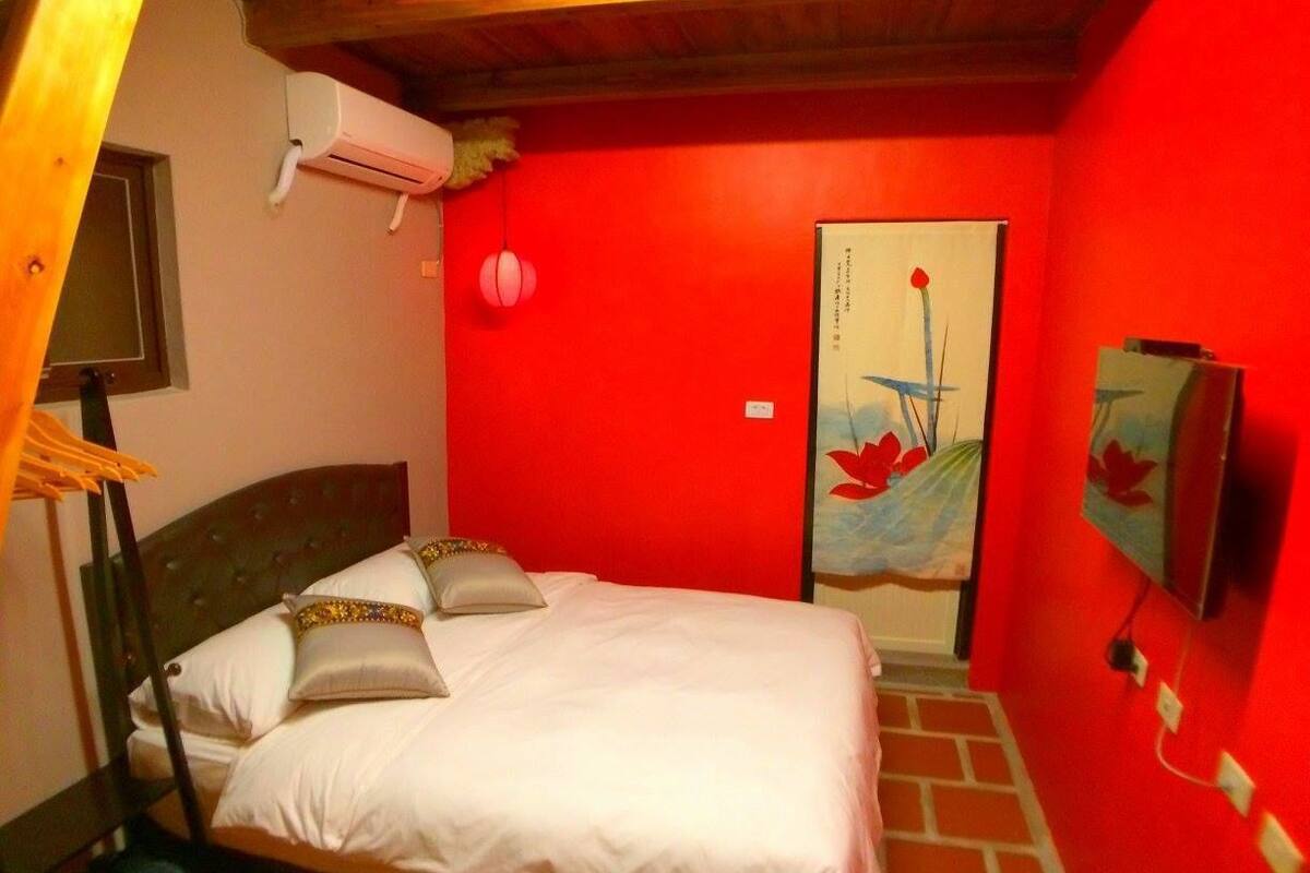 A hotel room with a bed, a red wall, and a painting of a boat.