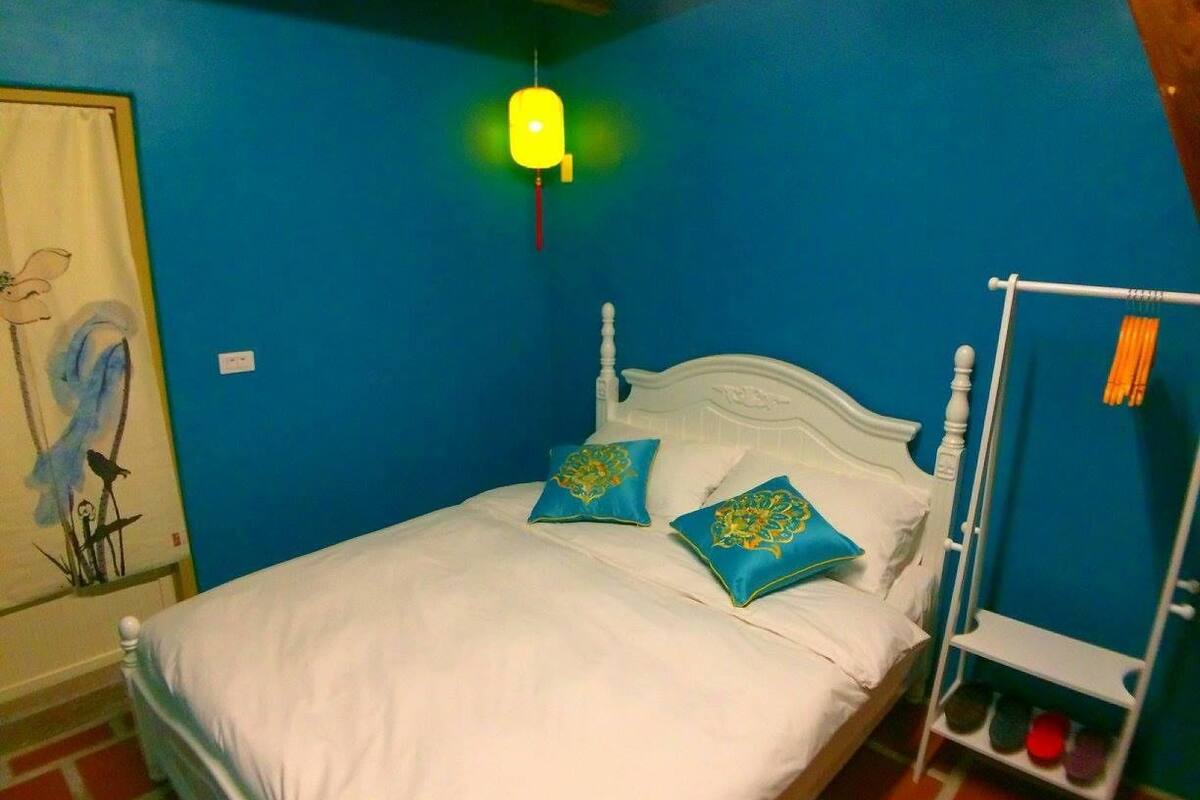 A bed with white bedding and two blue pillows, a white headboard, a blue wall, a yellow hanging light, and a shoe rack with shoes.