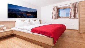 Une chambre moderne avec un lit en bois, une literie blanche, un plaid rouge, une table de chevet, une lampe, une fenêtre avec des rideaux et un tableau représentant un paysage urbain accroché au mur.
