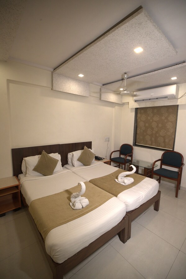 A hotel room with two beds, a desk, two chairs, a ceiling fan, and a window with a curtain. - Hotel The Excellency (Surat)