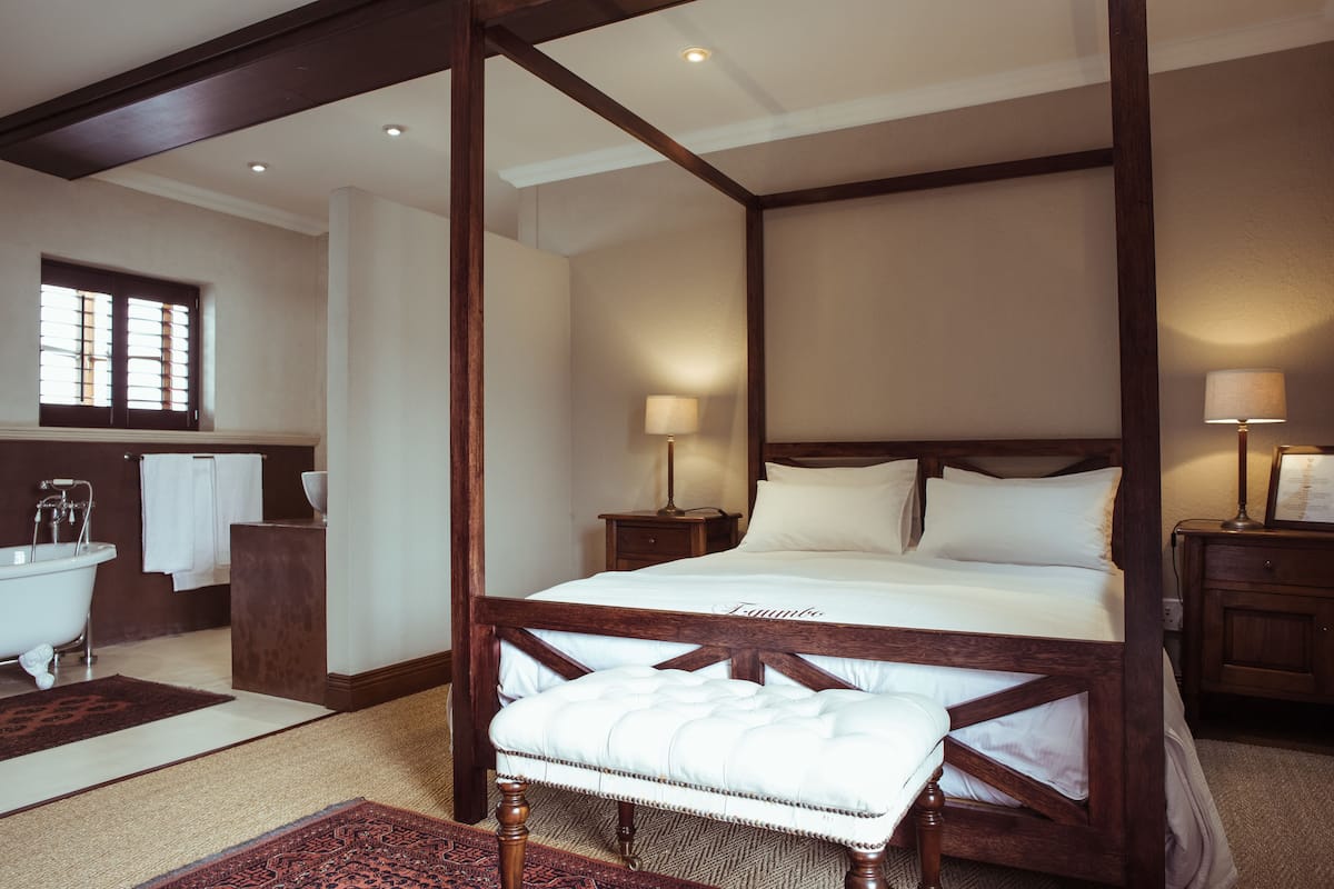 Une chambre spacieuse avec un grand lit, une tête de lit en bois, deux tables de chevet avec des lampes et une vue sur une salle de bain équipée d’une baignoire et d’un porte-serviettes.