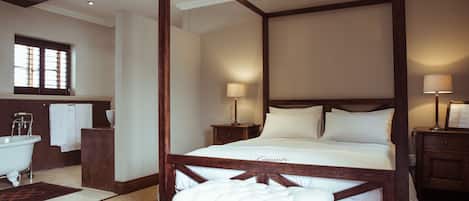 A spacious bedroom with a large bed, wooden headboard, two bedside tables with lamps, and a view of a bathroom with a bathtub and towel rack.