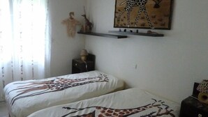 A bedroom with two beds featuring giraffe-themed bedding, a wall-mounted shelf with decorative items, and a window with sheer curtains. - nicely furnished and comfortable, close to the beach (Hourtin-Plage)