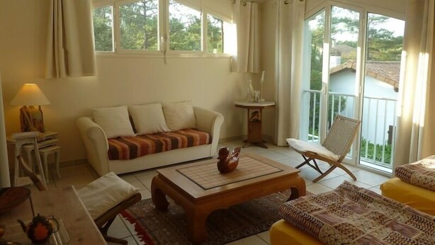 A spacious living room with a sofa, coffee table, and a chair. There are large windows and a balcony visible. - nicely furnished and comfortable, close to the beach (Hourtin-Plage)