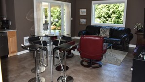 A modern living room with a glass dining table, red and black bar stools, a black leather sofa, and a large window with a view of greenery. - Executive Suite with commanding views - Claudette Ct. (Victoria)