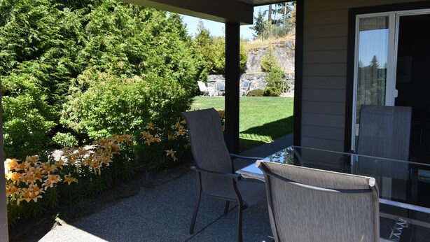 A covered patio with two chairs and a table, overlooking a garden with trees and a lawn. - Executive Suite with commanding views - Claudette Ct. (Victoria)
