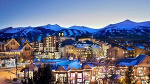 A town with snow-covered mountains in the background, featuring a mix of traditional and modern buildings with illuminated windows and signs. - Spring Break, Ski-in/out, 5-star, 2BR, sleeps 8 (Breckenridge)