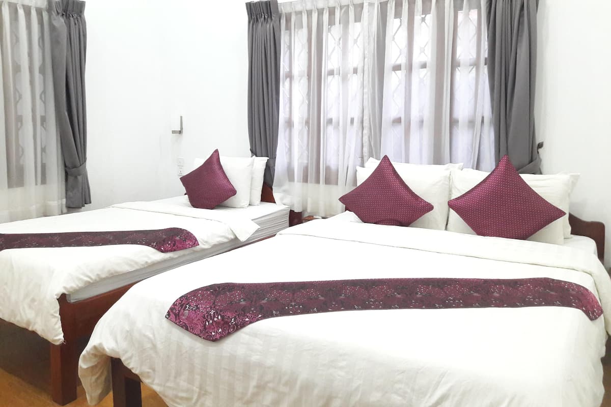 Habitación de hotel con dos camas individuales, cada una con ropa blanca y una manta morada. El suelo es de madera y las paredes son blancas.