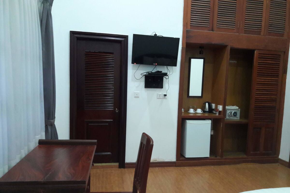 Una habitación con escritorio de madera, una silla, un refrigerador pequeño, un televisor montado en la pared y un armario de madera con espejo.