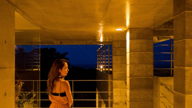 A woman in a long dress standing in a corridor with a view of a building in the background. - The:MOVES-Musée Villa- (Nakijin)