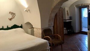 Ein Zimmer mit einem Bett, einem Sessel und einem Schreibtisch. Eine Wand ist mit einem dekorativen Element und einer Leuchte versehen. Ein Bogen führt in einen weiteren Raum.