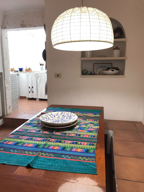 Comedor con una mesa sobre un tapete colorido, una lámpara colgante y vista de la cocina al fondo.