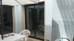 Una stanza piccola e pulita con un tavolo e sedie bianche, una porta vetrata che dà su un balcone e un bagno con lavandino e pareti piastrellate.