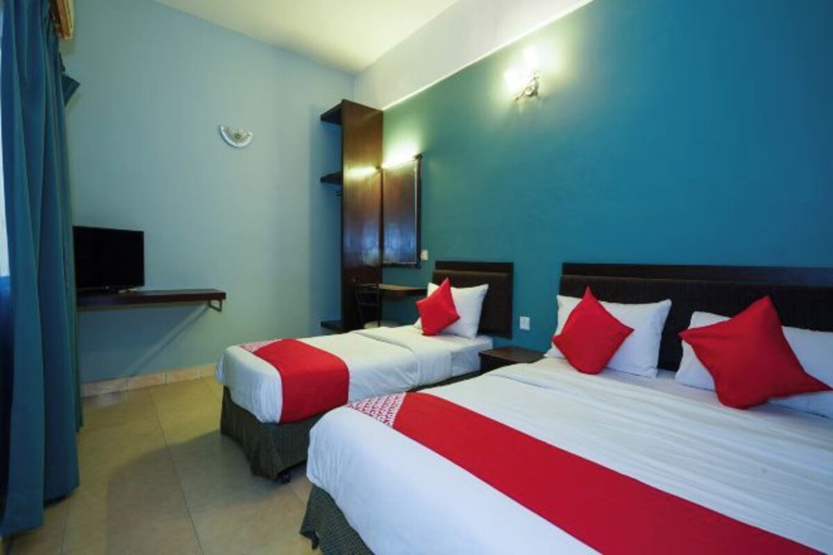 A hotel room with two beds, each with white bedding and red accents, a small desk, a television, and a wall-mounted light fixture.