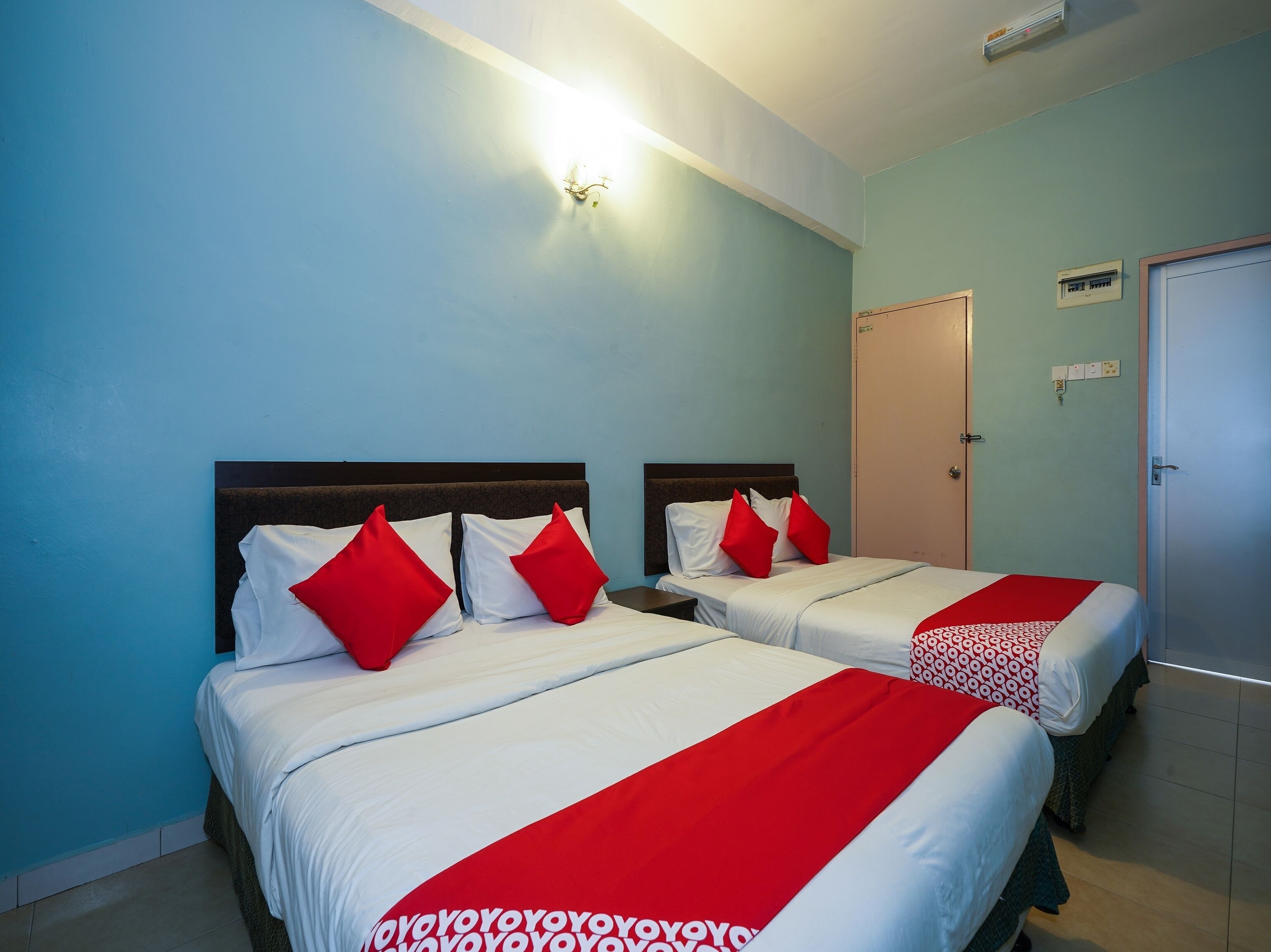 Een hotelkamer met twee bedden, elk met witte lakens en rode kussens, een lichtblauwe muur en een deur met een klein raam.