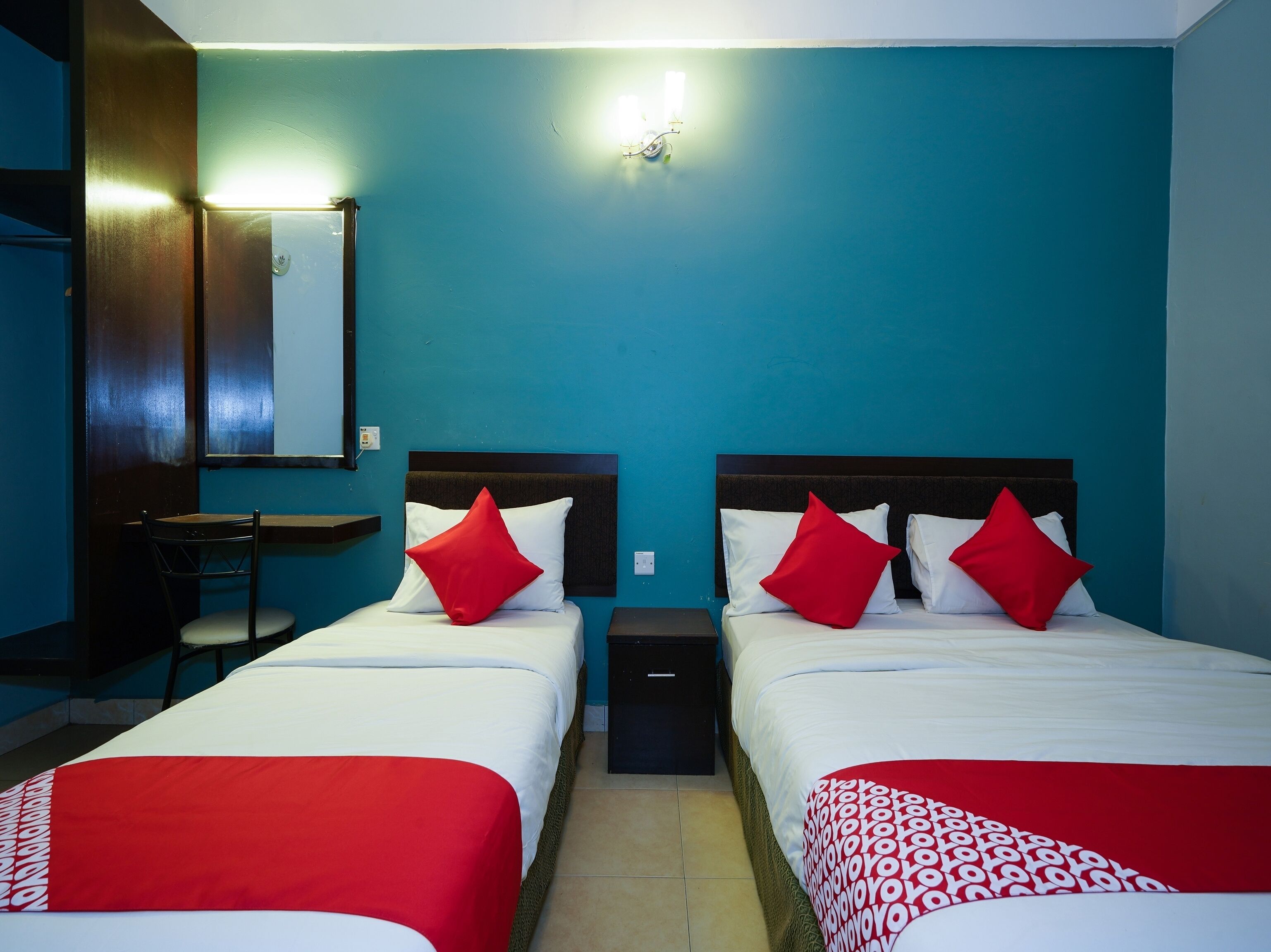 A hotel room with two beds, each with red and white bedding, a desk with a chair, and a wall-mounted light fixture.