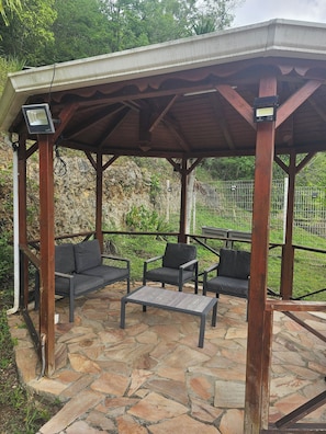 A covered outdoor seating area with stone flooring and wooden furniture.