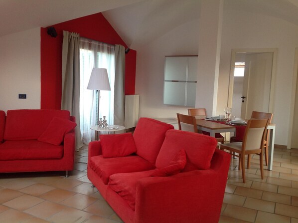 A living room with red sofas, a dining table with chairs, a lamp, and a TV mounted on the wall. - At the number in Viverone (Viverone)