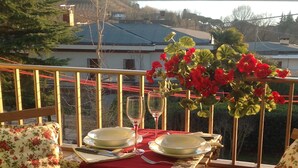 Une table dressée pour deux personnes, avec une nappe rouge, deux verres à vin et un vase de fleurs rouges, sur un balcon donnant sur un lac et des collines.