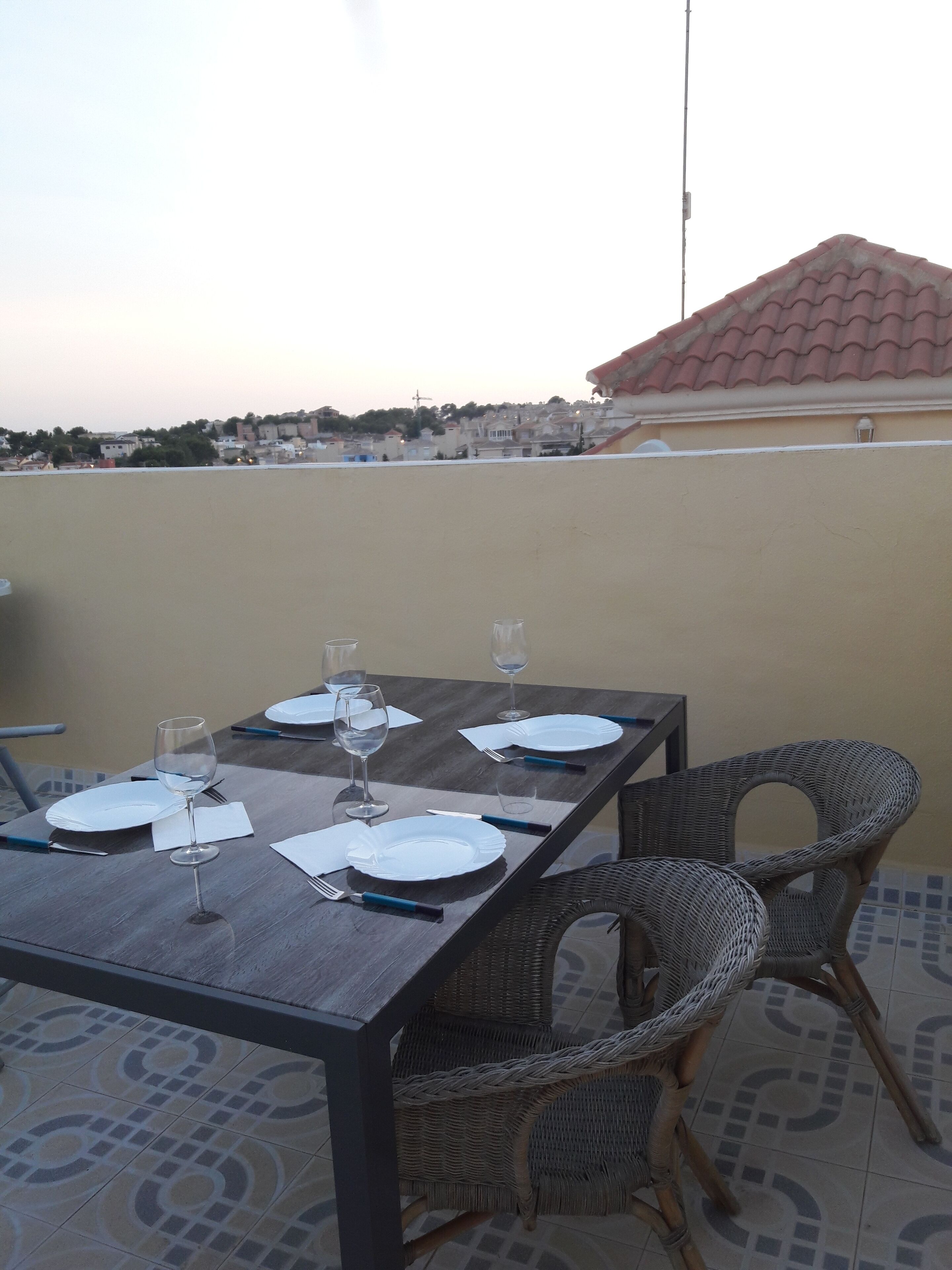 Mesa de jantar posta para quatro, com cadeiras, piso de azulejos e vista para uma área residencial ao fundo.