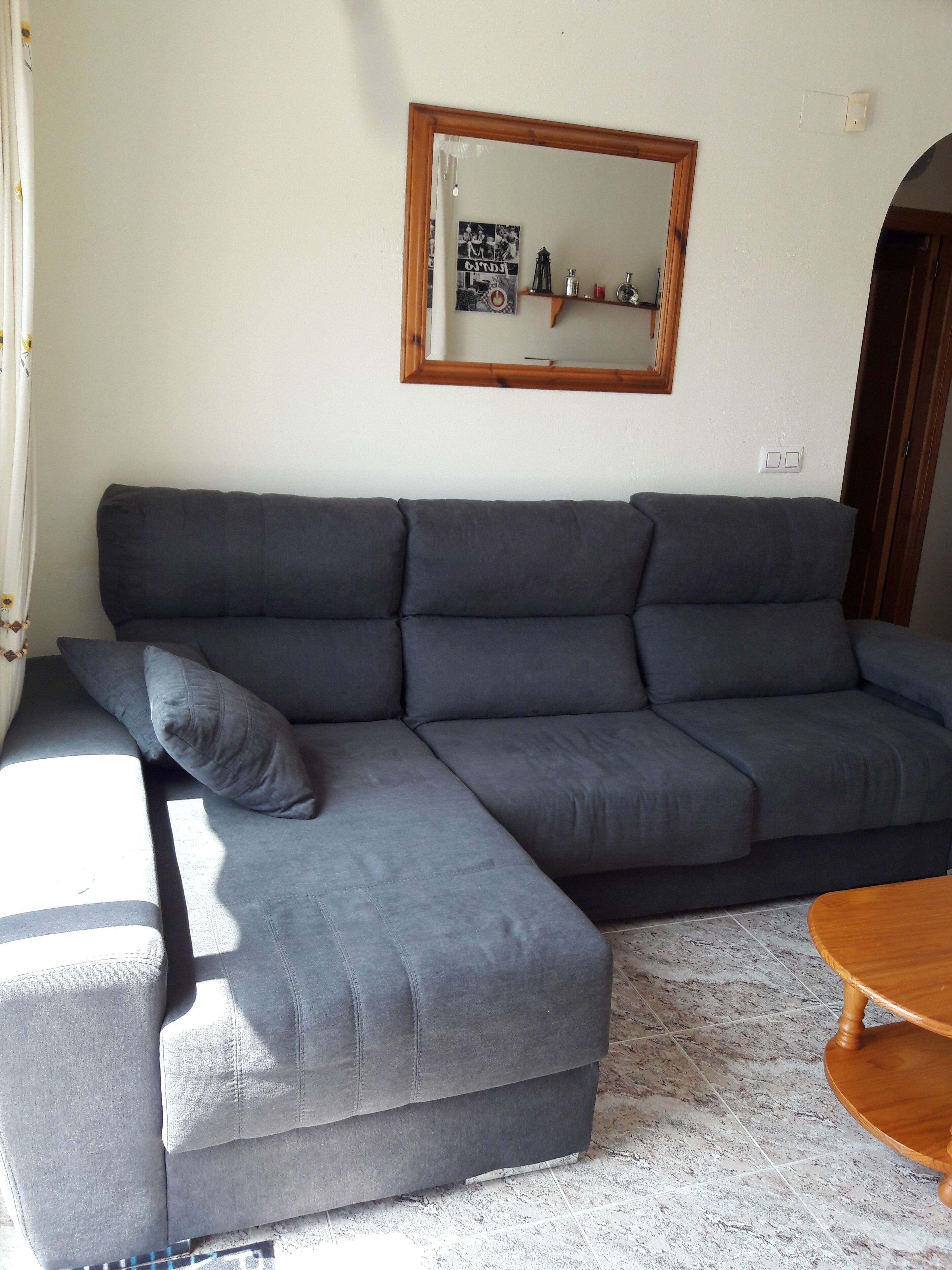 A grey sectional sofa with a wooden frame, a small wooden table, and a wall-mounted mirror.