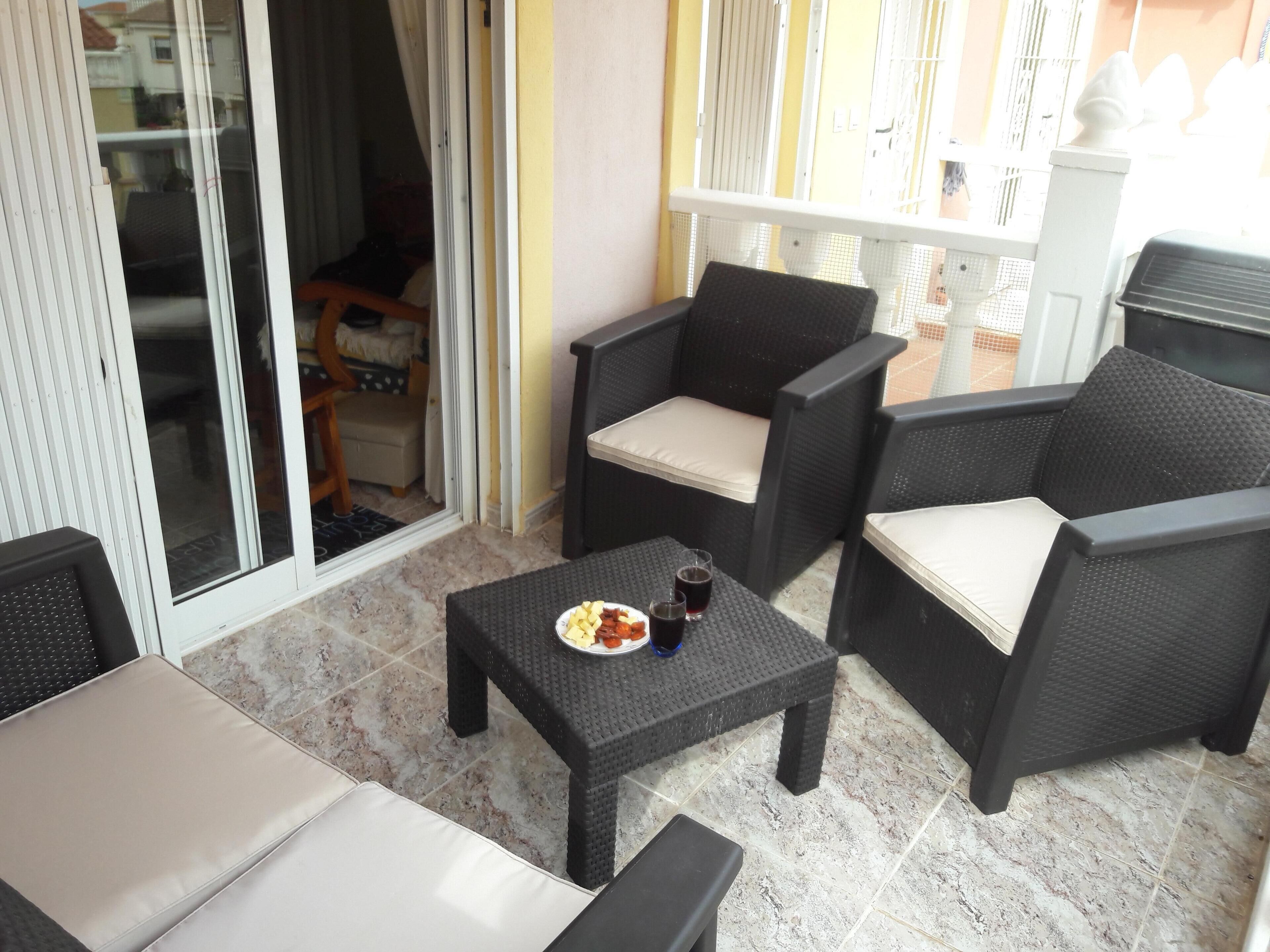 Een terras met rieten meubilair, een kleine tafel met eten en drinken, en een schuifpui die leidt naar een binnenruimte.