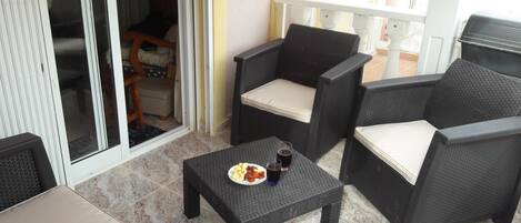 Een terras met rieten meubilair, een kleine tafel met eten en drinken, en een schuifpui die leidt naar een binnenruimte.