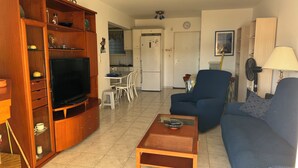 A living room with a TV, sofa, and coffee table. There is a kitchen in the background with a fridge and a dining table.