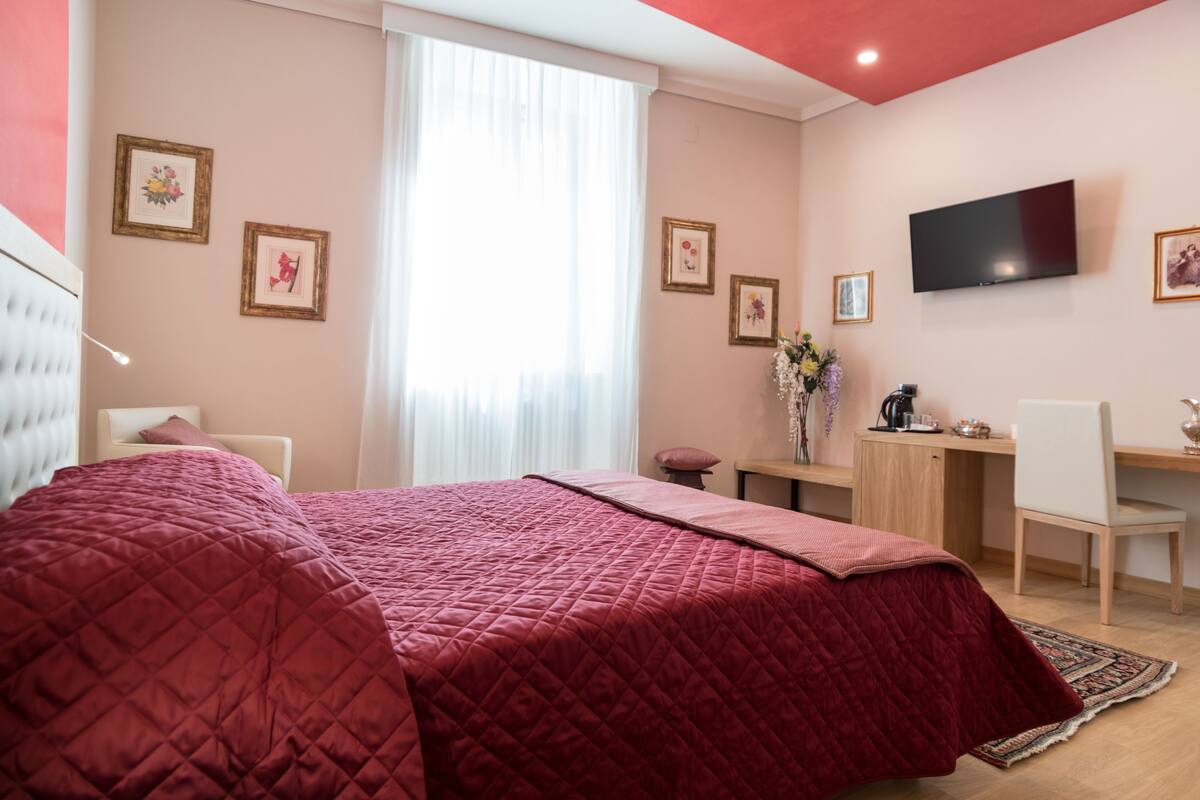 A hotel room with a large bed, a desk, a chair, a TV, and framed pictures on the wall.