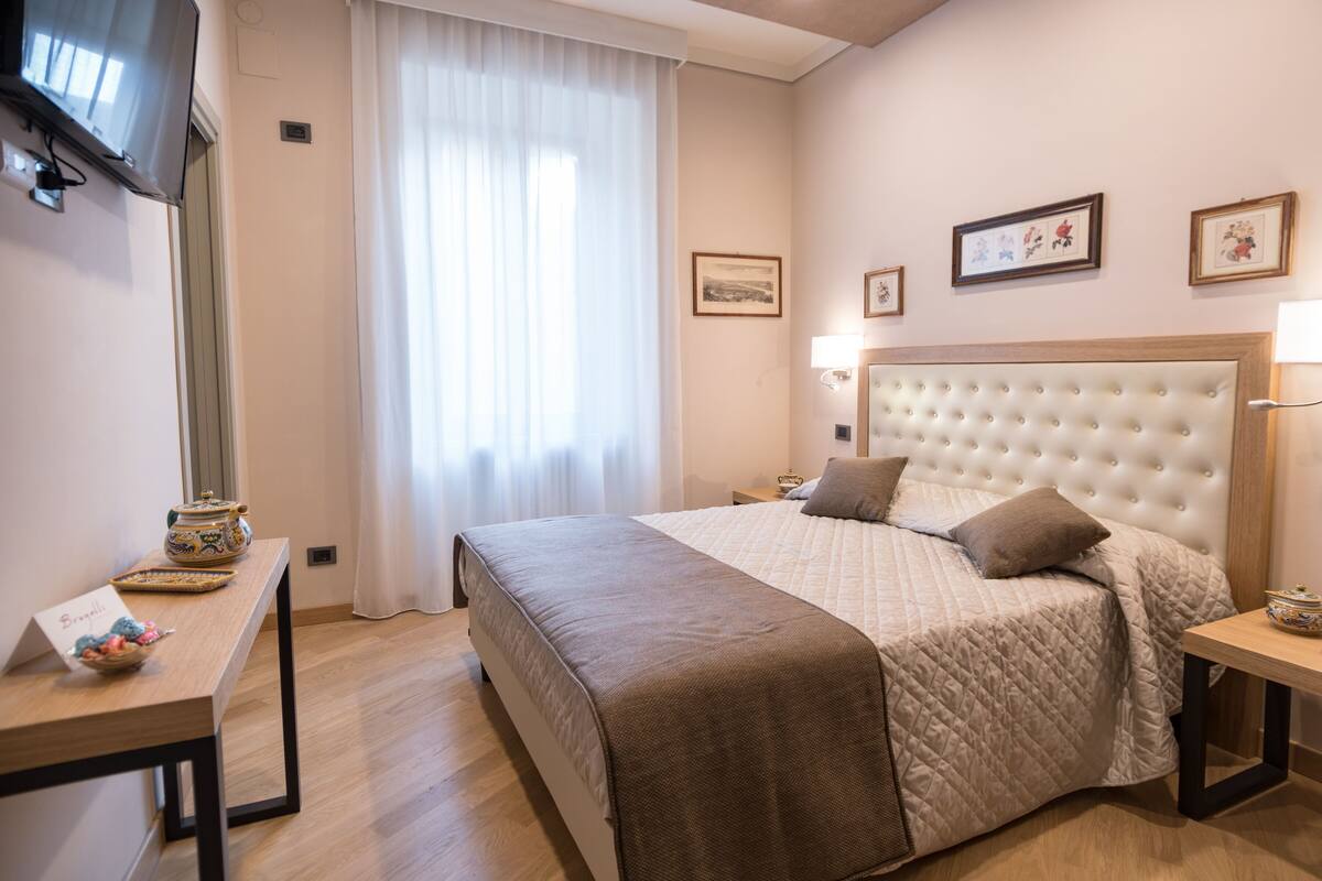 A hotel room with a large bed, a bedside table, a TV mounted on the wall, and a window with curtains.