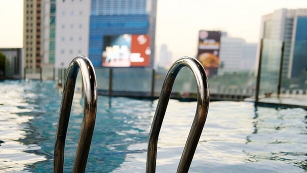 A poolside view with metal pool ladders, a cityscape in the background, and a building with a blue and white facade. - GP Plaza Apartment (Jakarta)