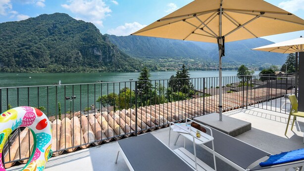 A balcony with a view of a lake and mountains, featuring lounge chairs, a table, and an umbrella. - Directly on the lake with two bedrooms (Idro)