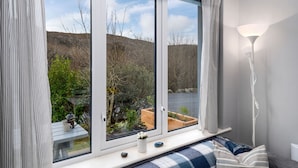 A room with a large window, a bedside table with a mug, a lamp, and a view of a garden and hills. - Cottage 315 -  Clifden - sleeps 4 guests  in 2 bedrooms (Clifden)