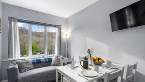 A modern dining area with a white table, chairs, and a sofa. A large window provides natural light, and a flat-screen TV is mounted on the wall. - Cottage 315 -  Clifden - sleeps 4 guests  in 2 bedrooms (Clifden)