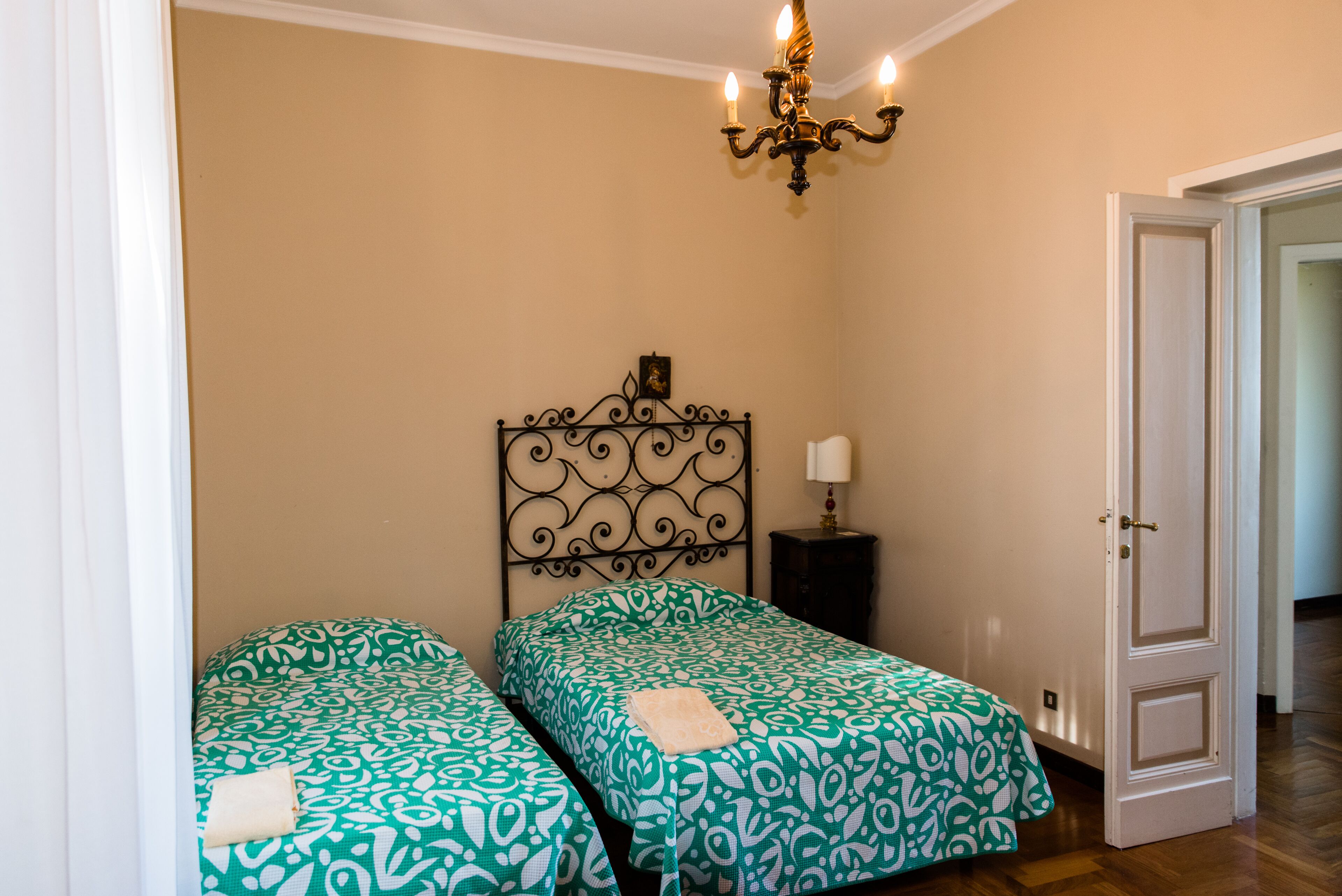 Quarto com duas camas individuais, um candelabro, uma mesa de cabeceira com abajur e uma porta para outro cômodo.