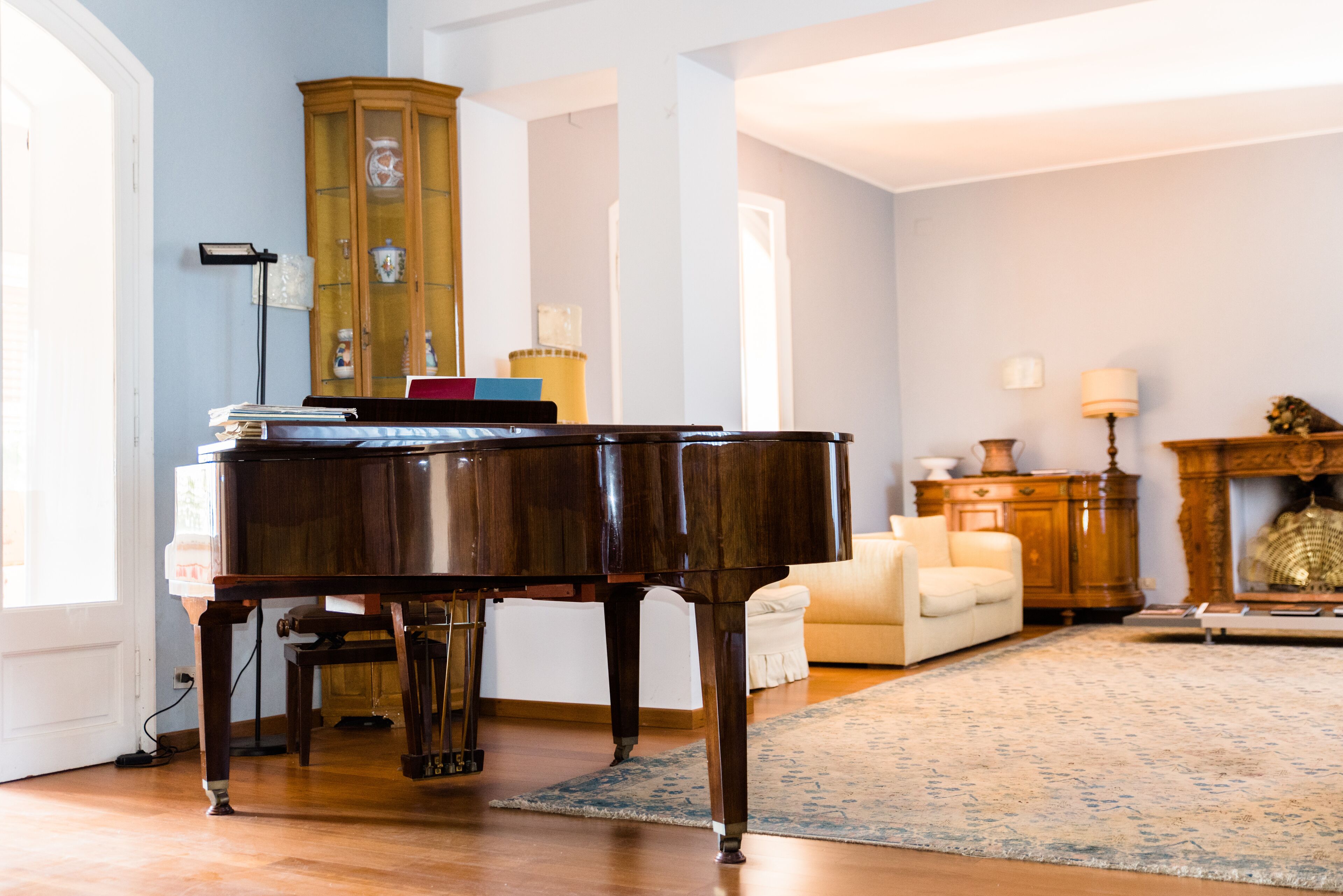Un piano de cola en un salón luminoso con techos altos, un aparador de madera con puertas de cristal y un rincón de descanso acogedor con un sofá y una lámpara.