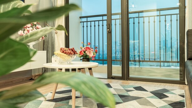 Un salon moderne avec un balcon, une table ronde ornée de fleurs et un tapis à motifs géométriques.