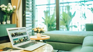 Un ordinateur portable est posé sur une table ronde, avec vue sur un balcon et sur la verdure à l’extérieur.