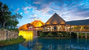 Un resort con una estructura de techo de paja, un edificio de paredes de cristal y un puente sobre una masa de agua reflectante.