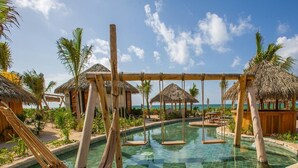 Un área de piscina con un conjunto de columpios, palmeras y estructuras de techo de paja.