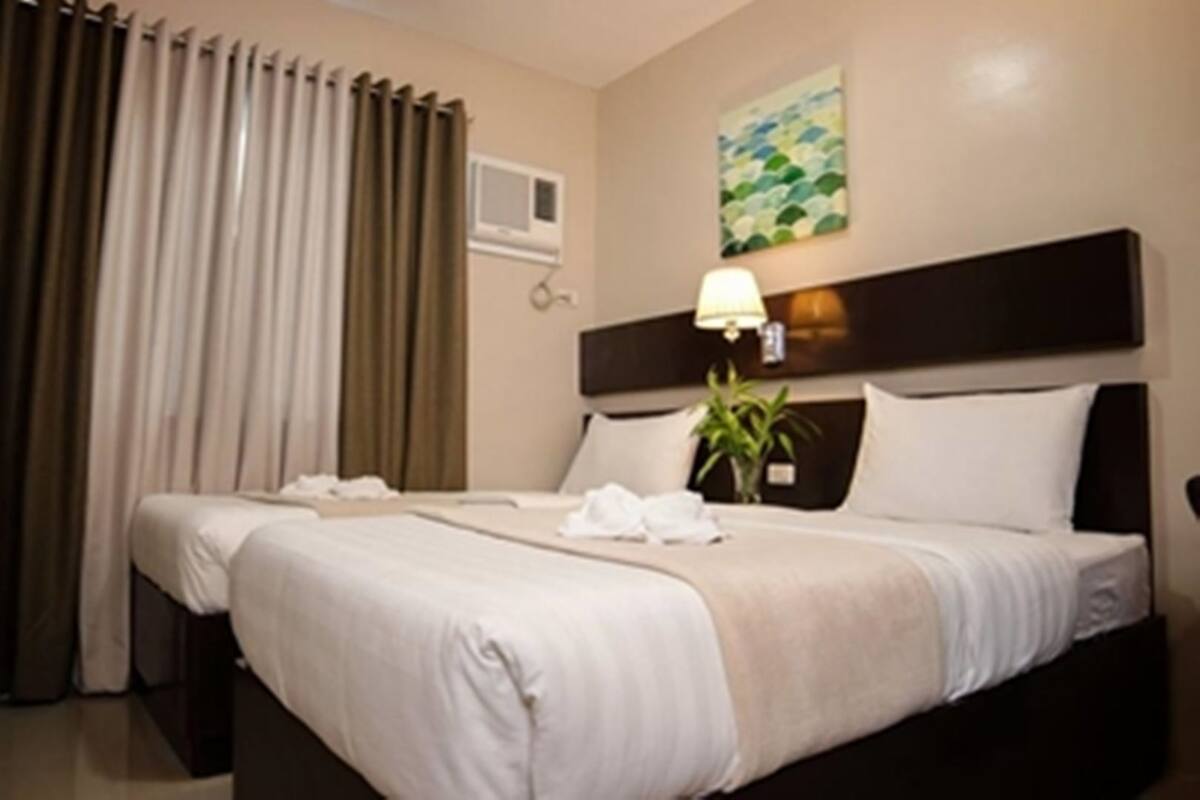 A hotel room with two beds, a large window with curtains, a wall-mounted air conditioner, a painting, and a bedside lamp.