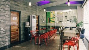 Un espace repas moderne avec une longue table et des chaises rouges, un mur en pierre et une cuisine visible à travers une porte ouverte.