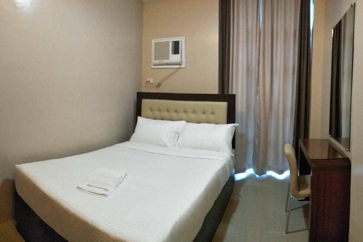 A hotel room with a bed, a bedside table, a chair, a window with curtains, and an air conditioning unit on the wall.