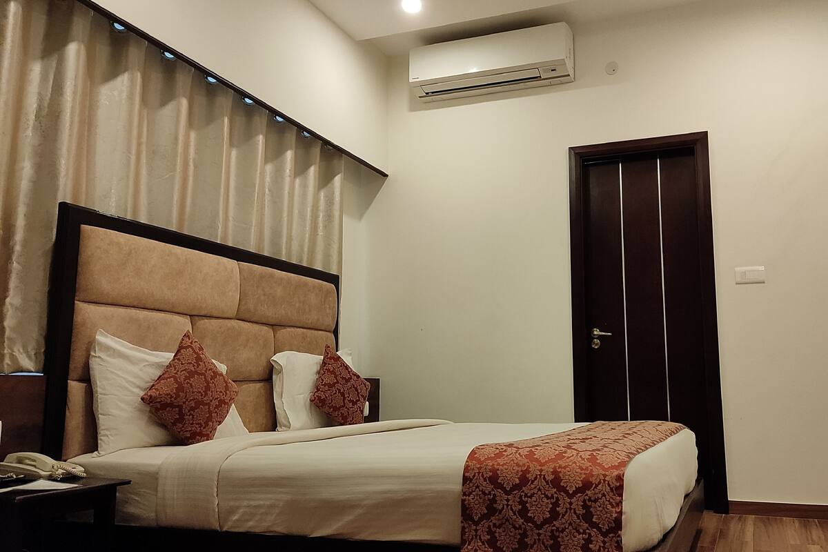 A hotel room with a bed, a nightstand, an air conditioning unit, a door, and a television mounted on the wall.