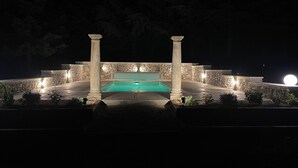 Vue nocturne d’un espace piscine avec des murs et des colonnes en pierre, entouré de plantes et éclairé par un éclairage d’ambiance.
