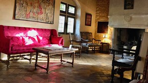 Une pièce au décor traditionnel, avec un canapé rouge, une table en bois et une cheminée. Un grand tableau orne le mur, et un mur de pierres apparentes se trouve au fond de la pièce.