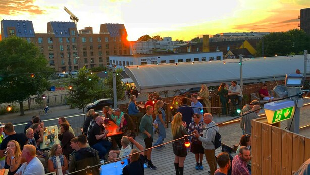 Un bar en la azotea con gente conversando, un edificio grande al fondo y un cielo al atardecer.