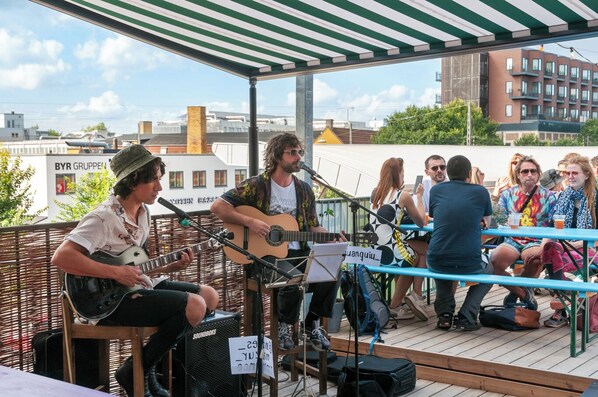 A band performing on a wooden deck with a green and white striped canopy, surrounded by seated audience. - Urban Camper Hostel (Copenhagen)