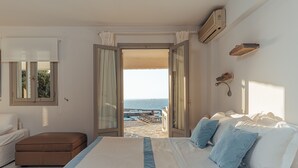 A bedroom with a large bed, a view of the sea through an open door, a small table with chairs outside, and a white wall with a mirror and air conditioning unit. - TERRA retreat in Paros, with prIvate pool, stunning sea view, near sandy beaches (PARIKIA PAROS)