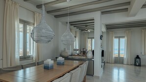 A spacious dining area with a wooden table and chairs, two hanging light fixtures, and a view of the sea through large windows. - TERRA retreat in Paros, with prIvate pool, stunning sea view, near sandy beaches (PARIKIA PAROS)